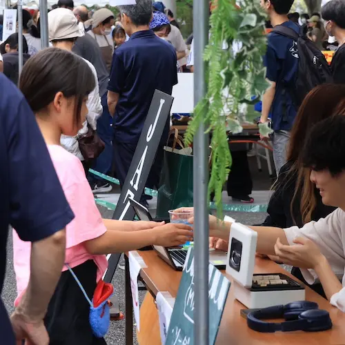 屋外の販売ブースで、ピンクのTシャツを着た来場者がプラスチックカップを受け取り、スタッフがノートPCで対応している